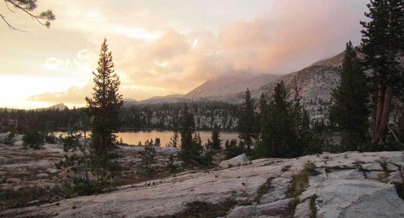 Under a pink, yellow and purple sky, a lake is nestled among mountains and evergreen trees. 